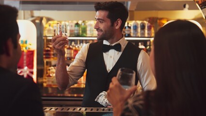 Focusing smiling smart bartender serving gin fizz or tonic soda at luxurious bar, engaging lively conversation with VIP customer couple at back view in atmosphere at lighting warm nightclub. Vinosity.