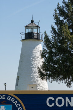 Havre de Grace, Maryland US - April 12, 2023: Concord Point Lighthouse seen from the street, with a tree and wooden sign from the park it sits in, located in the foreground