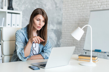 Young attractive casually dressed woman teacher or tutor having online lesson talking to her student via laptop computer while sitting in light colored modern office or classroom