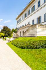 Obraz premium Visitors stroll through the lush gardens of Villa Melzi in Bellagio, enjoying the view of the neoclassical architecture against the serene backdrop of Lake Como on a sunny day.