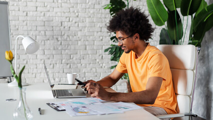 Portrait of a young handsome casually dressed African American business man working in his start up project using mobile phone and laptop computer while sitting in modern light colored office