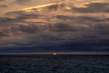 The sun is slowly setting over the vast ocean under a cloudy sky