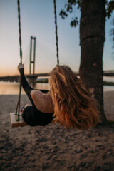 Beautiful young teenage girl rides on a swing near the water in the city at sunset