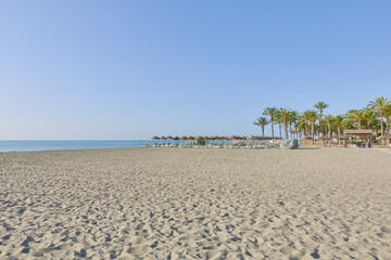 Beach of Los Alamos in Torremolinos with a palm grove on the sand, sunbeds, straw umbrellas in the seashore. In Malaga, Andalusia, Spain