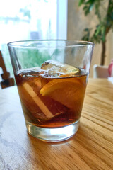 Closeup of cold liquid, lemon slice and ice cubes in glass with vermouth alcoholic drink on wooden table in cozy restaurant or home with backlight