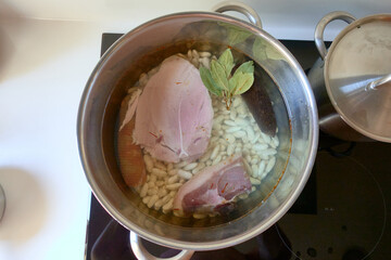 A stainless steel pot on a hob containing water, white beans, piece of ham, chorizo, blood sausage,...