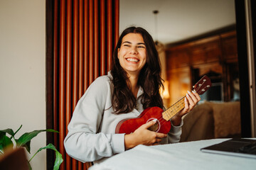 Young woman practicing ukulele guitar while reading notes on laptop	