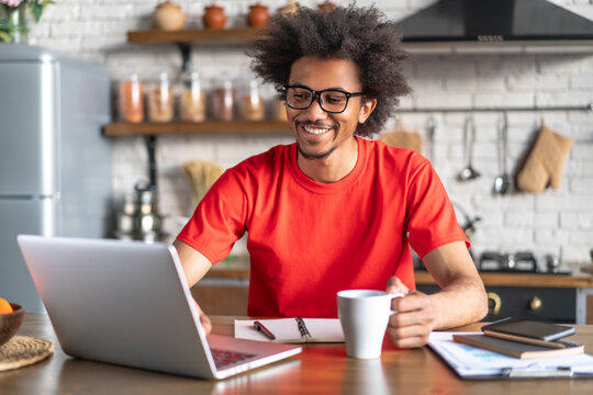 Joyful young attractive African American man working from home using his laptop computer, drinking coffee while sitting in modent light colored loft interior kitchen