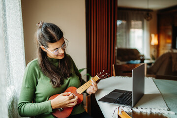 Young woman practicing ukulele guitar while reading notes on laptop	