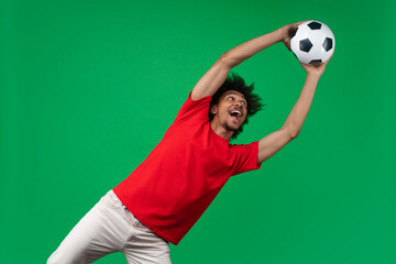 Studio portrait of excited joyful young African American football fan man in basic red t-shirt supporting favorite team catching soccer ball isolated over green background