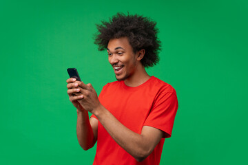 Studio portrait of casually drsssed excited joyful surprised young African American man reading message on his mobile phone isolated on green background