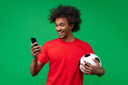 Handsome smiling African American young man holding soccer ball and watching online broadcast on his mobile phone cheering for favourite team, making bets online and waiting for winning results