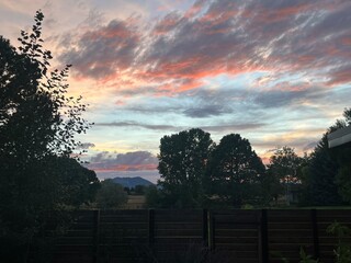 The sky is filled with vivid colors as the sun sets behind the mountains, casting a warm glow over the trees and the tranquil rural landscape in the early evening.