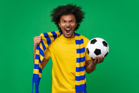 Studio shot of euphoric screaming soccer fan man wearing yellow-blue scarf posing with a ball in hand over green background cheering up for his favorite team