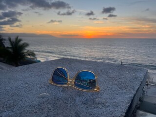 Golden sunglasses resting on a concrete ledge overlooking a vibrant sunset over the ocean