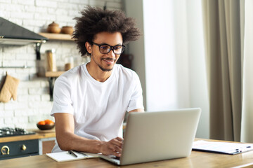 Casually dressed African American entrepreneur working with laptop computer remotely while sitting at wooden table in the light colored loft interior kitchen