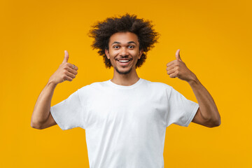 Portrait of happy smiling handsome young African American man in basic white t-shirt posing in studio showing thumbs up gesture over bright colored orange yellow background