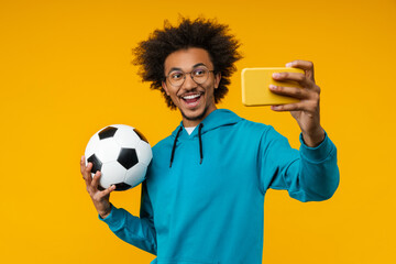 Studio portrait of happy smiling football supporter taking selfie on his mobile phone with soccer ball in hands, isolated over bright colored orange yellow background
