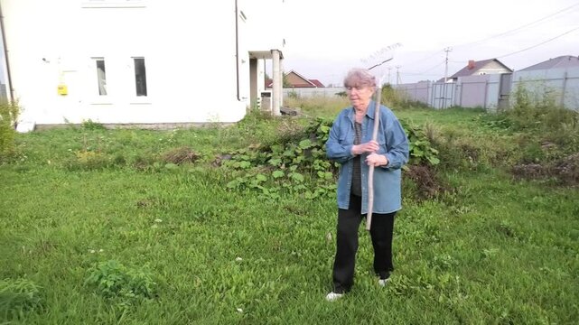 An elderly woman walks through the grass on a summer evening, carrying a rake on her left shoulder
