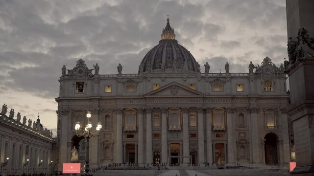 Slow-motion forward footage of Saint Peter&rsquo;s Square, Vatican City, Rome, Italy during the blue hour with glowing lamps and a cloudy sky while people appreciate highlighting its majestic architecture