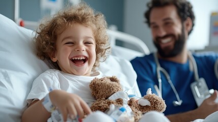 A happy child with curly hair is laughing while lying in a hospital bed, holding a teddy bear, with a male medical professional by their side.