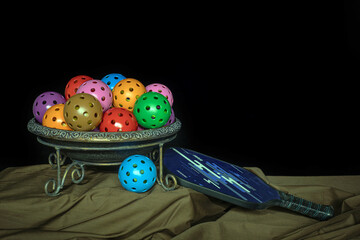 Bowl of colorful Pickleballs with Pickleball paddle on a black background. Pickleball stilll life photo.