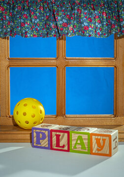 Yellow Pickleball in window setting with curtain and blue background.  Block letters "PLAY" in the forground.