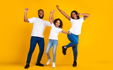 Having Fun. Full body length of excited young black family of three dancing and fooling around. Happy African American man, woman and girl enjoying favorite music together on yellow studio background