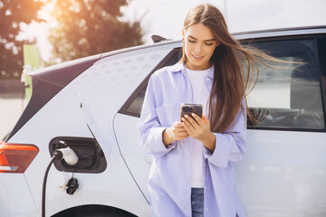 Young Woman Charging Electric Car While Using Smartphone at EV Charging Station on a Sunny Day