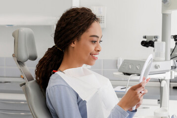 Beautiful black lady checking her white smile in mirror after stomatological treatment in modern...