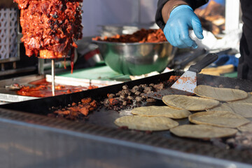 Street vendor grilling tortillas and chopped al pastor meat on a grill, in preparation of...