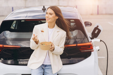 Naklejka premium Young Woman Enjoying Healthy Lunch While Charging Electric Car in Urban Parking Lot