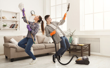 Young couple cleaning home, playing with mop and vacuum cleaner, having fun in living-room....
