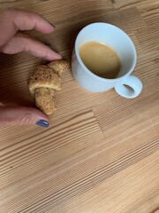 Minimalist wooden table with white cup of fresh espresso and female hand holding small croissant,...