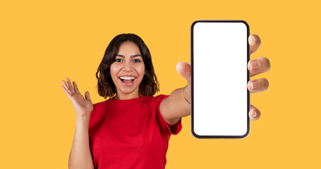 Emotional pretty young arab woman in red t-shirt showing brand new cell phone with white empty screen and gesturing over yellow studio background, online offer, mockup, panorama