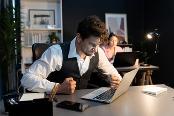 Sitting man with serious face thinking creative business project planning at night lighting time at modern office with blurry secretary checking paper report with folder background concept. Postulate.
