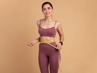 Weight Loss And Healthy Lifestyle Concept. Smiling slim woman measuring her waist with yellow tape over pastel studio background. Fit young lady in perfect shape, happy with her body