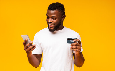 Online payment. Cheerful black man using credit card and smartphone for purchasing goods