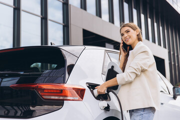 Woman Charging Electric Car While Talking On Phone Outside Office