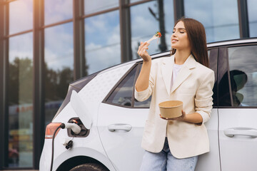 Naklejka premium Woman Enjoying Lunch Beside Charging Electric Car Outdoors