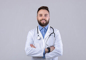 Portrait of positive male doctor posing with crossed arms and smiling at camera over grey studio background. Happy medical specialist in lab coat ready to fight viral diseases