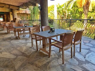 Empty wooden tables and chairs are set on a stone patio under a shaded area, overlooking a lush, green garden with palm trees. Concept of outdoor restaurant seating and tranquil dining.