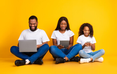 Family Gadgets. Portrait of happy African American parents and their little daughter holding and using different electronic devices while sitting on the floor over yellow studio background