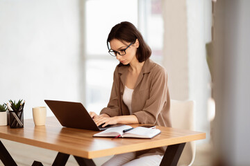 Home Office. Portrait of young female manager wearing specs and brown shirt using laptop, working remotely. Copy space