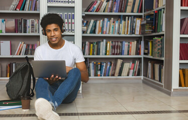 Young african american student with headset sitting on the floor, leaning on bookshelf and using laptop at library, free space
