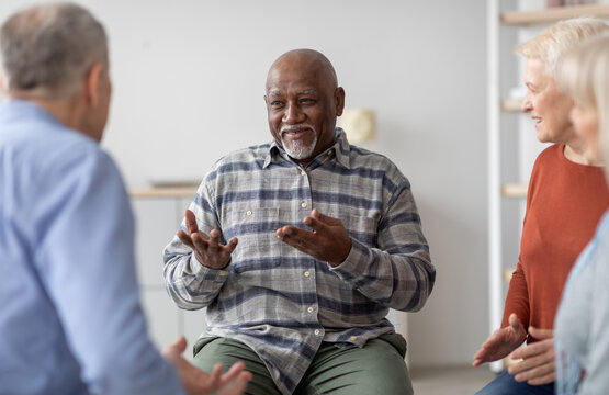 Cheerful african american elderly man having conversation with male coach while attending group therapy session with multiracial group of seniors, psychological support at nursing home concept