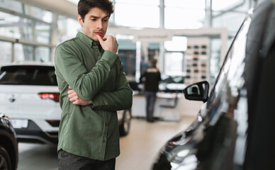 Pensive young guy thinking, making choice, having doubt about buying new car at dealership centre,...