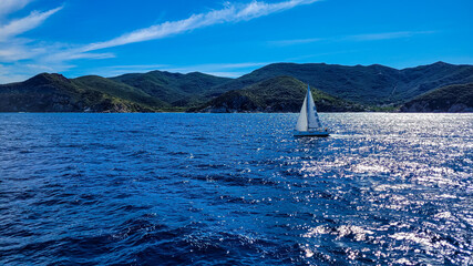 The sailboat with a white sail goes on the blue sea, lake or ocean. In the background are mountains covered with forests, a stone shore and a small town.