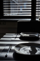 A dining table with a navy blue place mat, white plate, and silver cutlery. Sunlight through window blinds creates shadow patterns on the scene.