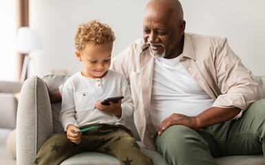 Black Grandfather And Little Grandson Using Cellphone Playing Mobile Game Together Sitting At Home....
